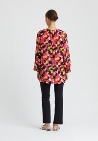 Bunte Bluse mit langen Ärmeln, die ein florales Muster aus Pink, Gelb und Schwarz auf einem pinken Hintergrund zeigt; kombiniert mit schwarzen Hosen.