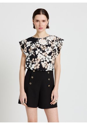 Femme portant un chemisier sans manches noir à fleurs et un short noir taille haute avec des boutons dorés, debout devant un fond gris clair.