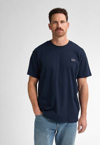 Námořnická modrá bavlněná tričko s kulatým výstřihem, krátkými rukávy a malou nášivkou na hrudi. Nosené s světle modrými džínami.