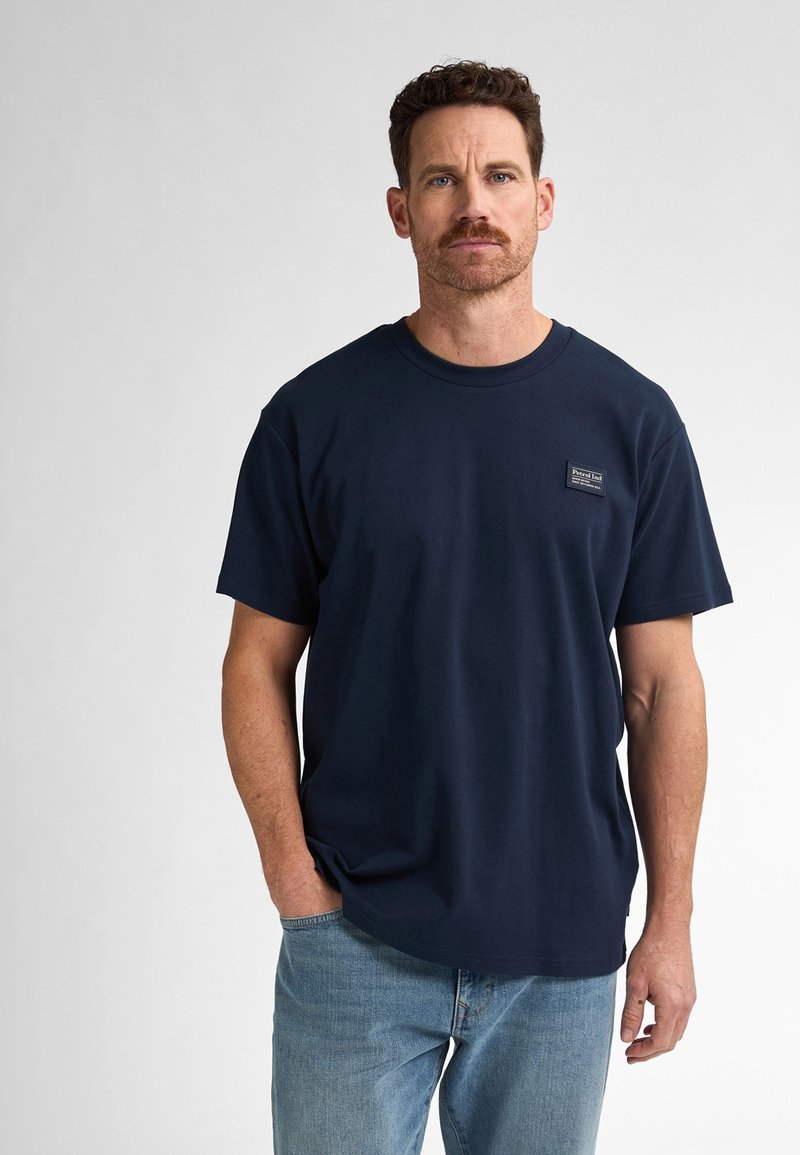 Námořnická modrá bavlněná tričko s kulatým výstřihem, krátkými rukávy a malou nášivkou na hrudi. Nosené s světle modrými džínami.