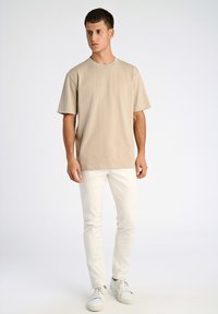 Bagefarvet t-shirt med korteærmer og rund halsudskæring, parret med tætsiddende hvide bukser og hvide sneakers, stående på en neutral baggrund.