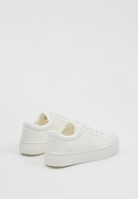 Zapatillas blancas con cordones, con un patrón floral texturizado, cuello acolchado y suela de goma blanca. La vista trasera muestra una forma redondeada y suave.