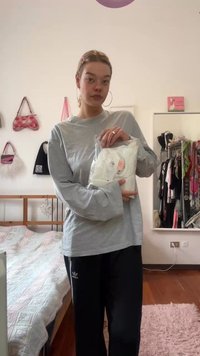 Siva majica dugih rukava drži se iznad bijelog pakiranja. Crne trenirke s tri pruge na nozi. Mekano posteljno rublje i stalak za odje�ću u pozadini.