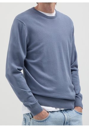 Mężczyzna w jasnoniebieskim swetrze z okrągłym dekoltem na białym t-shircie, z rękami w kieszeniach jasnoniebieskich dżinsów na jednolitym tle.