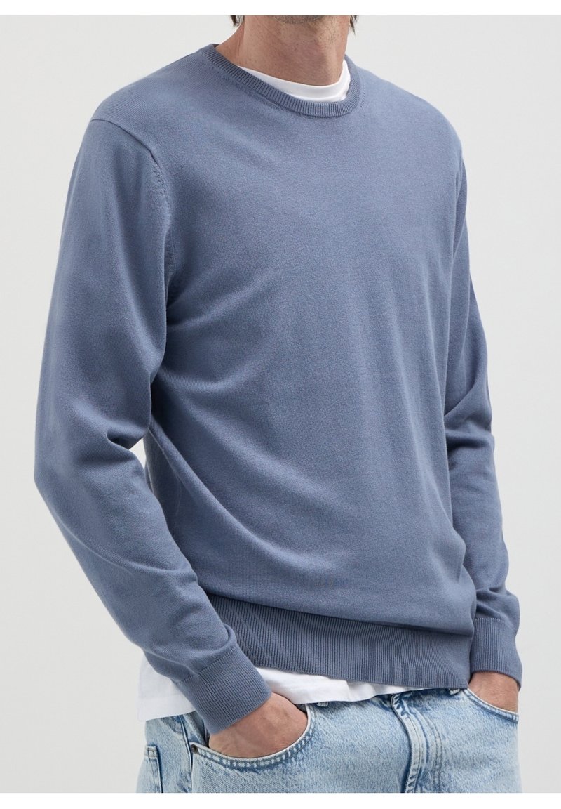 Homme portant un pull ras du cou bleu clair sur un t-shirt blanc, les mains dans les poches d'un jean bleu clair, sur un fond uni.