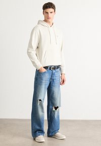 Junger Mann trägt einen cremefarbenen Kapuzenpullover, einen schwarzen Gürtel, ausgefranste lockere blaue Jeans und beige Turnschuhe, steht vor einem schlichten hellen Hintergrund.