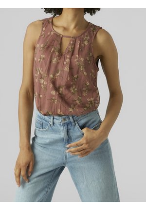 Femme portant un chemisier sans manches mauve à motifs floraux et un jean taille haute bleu clair, avec une main dans la poche, sur un fond neutre.