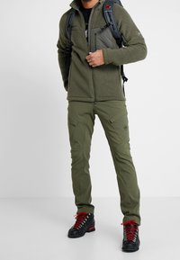 Olive zielona bluza polarowa o teksturowanej powierzchni, z kieszenią na piersi, w zestawie z pasującymi spodniami z kieszeniami cargo oraz czarnymi butami trekkingowymi z czerwonymi sznurówkami.