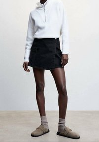 Suéter blanco con cremallera en el cuello, mini falda de mezclilla negra con bolsillo lateral, zapatos beige tipo slip-on combinados con calcetines grises, diseño minimalista.