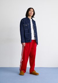 Denim krekls ar diviem krūšu kabatām un zilu diegu, kombinēts ar sarkaniem sporta biksēm ar baltām strīpām un brūniem klogiem uz zila grīdas.