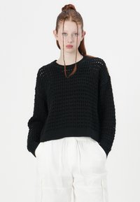 Pull en maille noire avec un motif texturé ouvert, des poignets et un ourlet côtelés. Porté avec un pantalon blanc à coupe décontractée pour un contraste.
