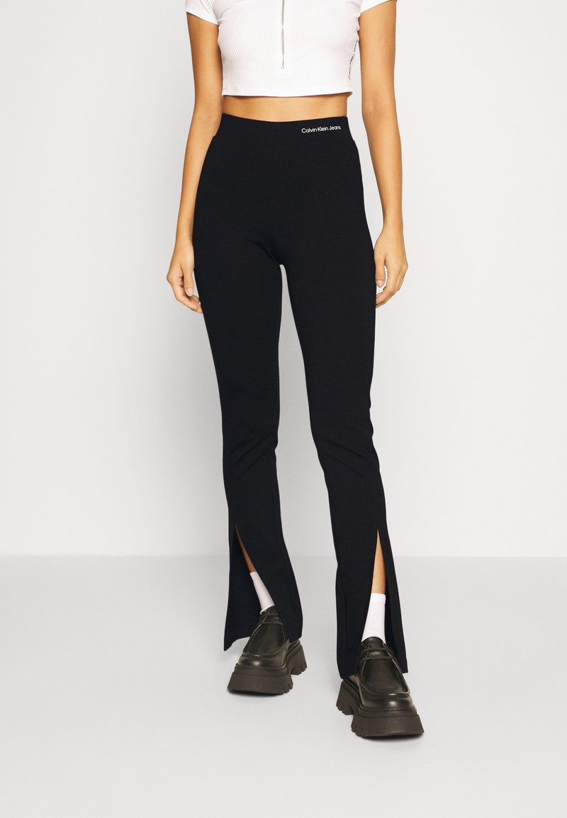 Zwarte geribbelde legging met zijsplitten, hoge tailleband met het Calvin Klein Jeans-logo, gecombineerd met donkere, stevige schoenen en witte sokken.