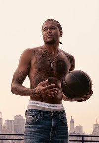 Athlet, der einen Basketball hält, trägt Blue Jeans und einen wei�ßen Calvin Klein Bund. Hat Tätowierungen an Armen und Oberkörper sowie geflochtenes Haar.