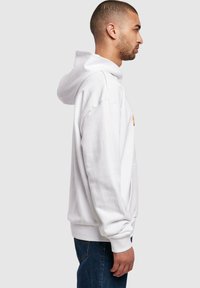 Hvid hætte sweatshirt med rummelig pasform, ribbede manchetter og kant, kengurulomme, båret over mørkeblå denimjeans. Vist fra siden.