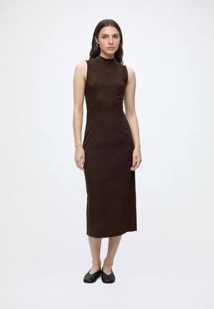 Femme portant une robe midi marron foncé sans manches à col montant, avec des chaussures plates noires, debout devant un fond blanc uni.