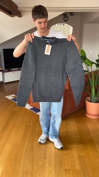 Mladý muž držící tmavě šedý svetr s visačkou uvnitř obývacího pokoje s dřevěnou podlahou a pokojovými rostlinami.