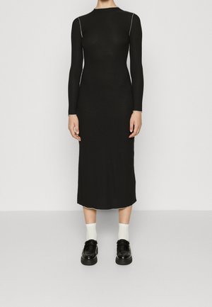 Kobieta w długiej czarnej dopasowanej sukience z białymi przeszyciami, białych skarpetkach i czarnych mokasynach, stojąca na jednolitym tle.