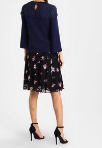 Femme portant un haut bleu marine avec col en dentelle, une jupe noire plissée à fleurs, et des sandales noires à talons hauts avec bride à la cheville, debout de dos.