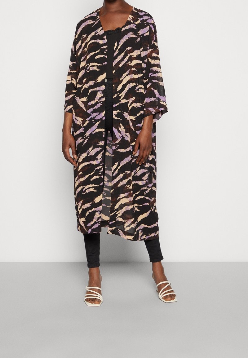 Personne portant un kimono ample, à motifs, jusqu'aux genoux, par-dessus des vêtements noirs et des sandales blanches à brides, debout devant un fond uni.