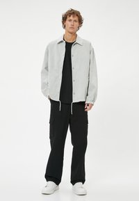 Harmaa denim-takki, jossa on napit, joustavat hihansuut ja säädettävä helma, yhdistettynä mustiin cargo-housuihin ja valkoisiin lenkkareihin.