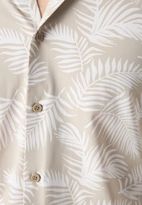 Lyhyellä hihalla oleva paita, joka on valmistettu teksturoidusta beigeistä kankaasta ja jossa on valkoinen lehtipalmukuosi sekä vaaleanruskeat napit edessä.