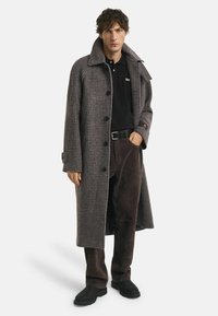 Man in een lange bruine geruite jas over een zwart poloshirt, donkere bruine broek, zwarte riem en zwarte schoenen, staande met één hand in zijn zak.