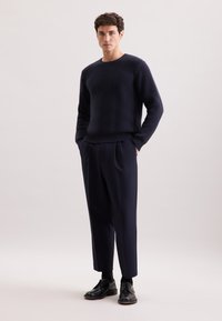 Granatowy sweter z prążkowanej dzianiny zestawiony z dopasowanymi ciemnymi spodniami i czarnymi sznurowanymi butami. Stylizacja charakteryzuje się luźnym krojem i jednolitą paletą kolorów.