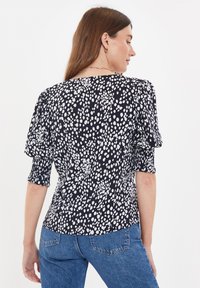 Sort bluse med hvidt prikmønster, korte pufærmer og rund halsudskæring. Bagsiden viser glat stof og tætsiddende design.