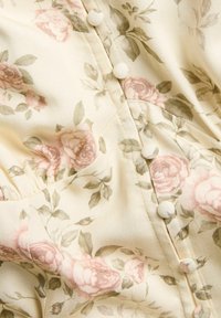 Blommigt tyg i krämfärgad med rosa och gröna rosor. Har runda knappar och rynkor längs sömmen för textur och form.