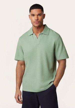 Man met kort haar, gekleed in een lichtgroen polo met korte mouwen en een donkere broek, staand met één hand in zijn zak tegen een beige achtergrond.