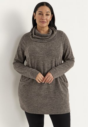 Kvinde med langt mørkt hår iført en brun tunika med lange ærmer og kittelhals samt sorte bukser, stående med hænderne foldet foran.