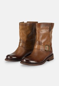 Bottes chevilles en cuir marron avec un design décontracté, présentant un détail de sangle à boucle et une pointe arrondie. La semelle est plate avec des accents texturés.