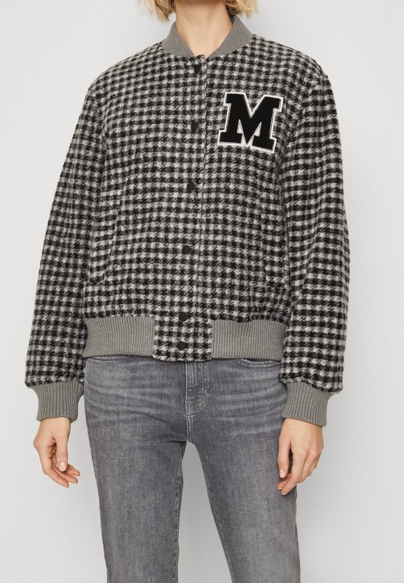 Personne portant une veste bomber en laine à carreaux noirs et blancs avec poignets et bas côtelés, et un grand écusson noir "M" sur la poitrine.