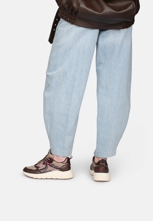 Person, die hellblaue, schmal zulaufende Hose und braune und beige Sneakers mit weißen Sohlen trägt, von hinten unterhalb der Taille gezeigt.
