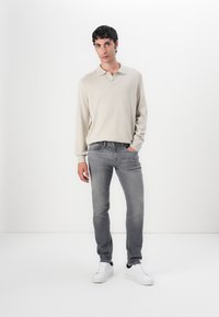 Világos bézs hosszúujjú póló ing, amelyet slim-fit szürke farmernadrággal és fehér sportcipővel párosítottak. A póló klasszikus gallérral rendelkezik és puha tapintású.
