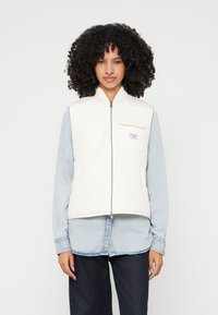 Bijela prsluk s quiltanom površinom, visokim ovratnikom, prednjim zatvaračem i džepom na grudima. Nosi se preko svijetloplavog traper košulje.