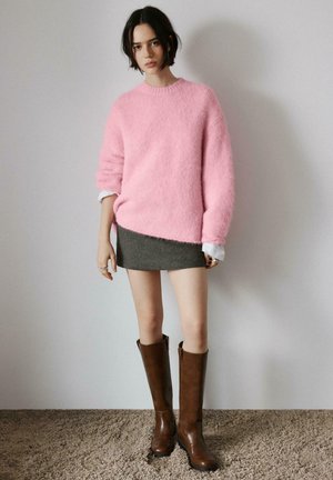 Jeune femme portant un pull rose duveteux, une mini-jupe grise et des bottes en cuir marron montant jusqu'aux genoux, debout sur un tapis devant un mur uni.