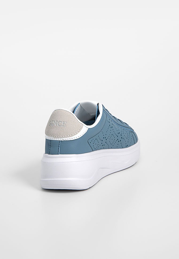 Zapatilla azul perforada con suela blanca, acento en el talón gris y textura de cuero suave. Presenta un sutil logo en la parte trasera.
