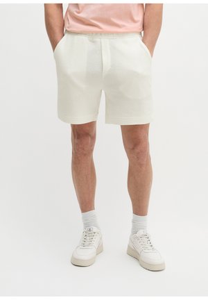 Man draagt off-white getextureerde korte broek met handen in de zakken, witte sokken en witte sneakers, staand tegen een effen achtergrond.