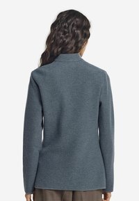 Kvinna med långt, vågigt mörkt hår, iklädd en texturerad blågrå långärmad jacka, stående vänd bort från kameran mot en enkel bakgrund.