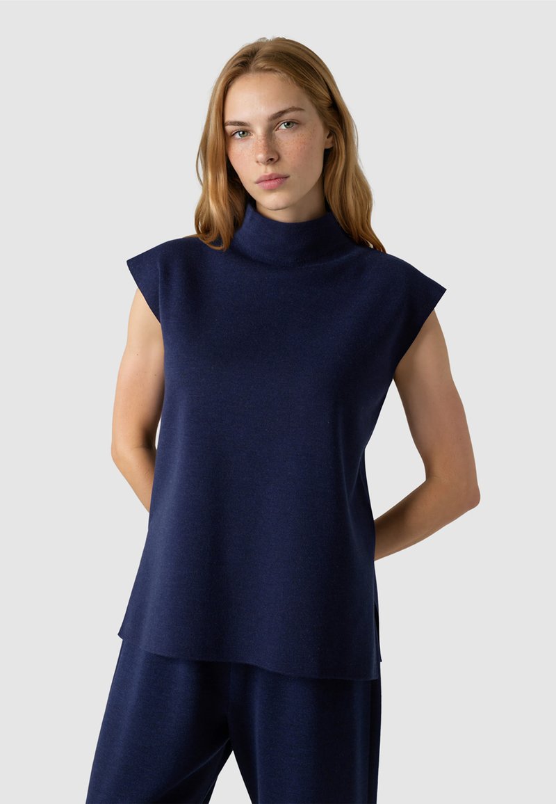 Jeune femme aux longs cheveux blonds portant un pull sans manches à col roulé bleu marine et un pantalon bleu marine assorti, debout les mains derrière le dos.