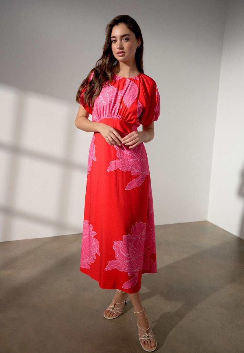 Woman standing indoors wearing a red midi dress with large pink floral prints and beige strappy sandals.