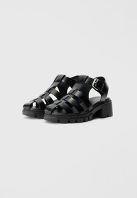 Svarte høye sandaler med en stroppedesign, laget av glatt syntetisk materiale. Har en kraftig såle og justerbar ankelspenne.