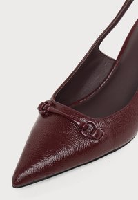 Chaussure slingback en cuir verni bordeaux à bout pointu avec une petite barre décorative et un détail anneau sur l’orteil.
