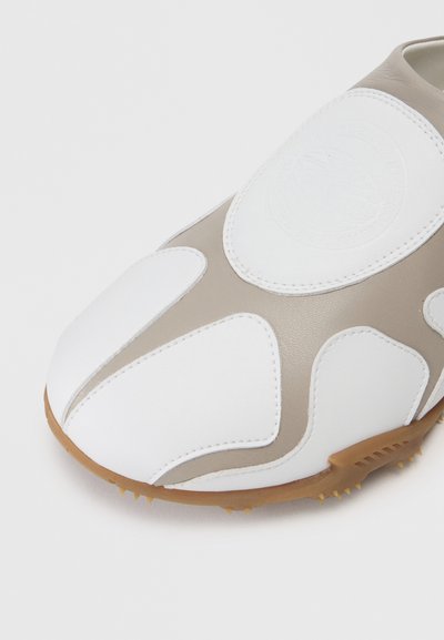 Chaussure de golf blanche et beige avec une tige en cuir lisse, un bout arrondi et des griffes en caoutchouc sur la semelle extérieure pour une meilleure adhérence. Présente un détail de logo.