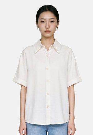 Donna con capelli scuri indossa una camicia bianca ampia a maniche corte con bottoni e jeans blu, in piedi davanti a uno sfondo chiaro e uniforme.