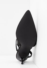 Chaussure noire à talon haut avec bout pointu incliné vers le haut, semelle texturée visible et talon aiguille fin sur un fond blanc d'angle.