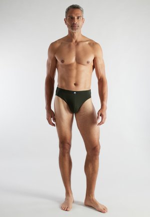 Fit adult man standing barefoot facing forward, wearing dark green briefs against plain light gray background.