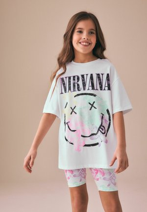 Uśmiechnięta dziewczyna z długimi włosami w białym, za dużym t-shircie Nirvany i pastelowych szortach w kwiaty na jednolitym tle.