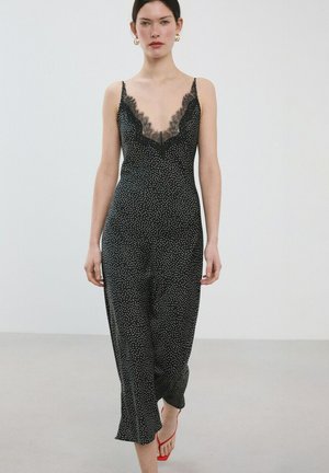 Mujer con vestido de tirantes negro con lunares y escote de encaje, y tacones rojos con tiras, de pie frente a una pared lisa y clara.